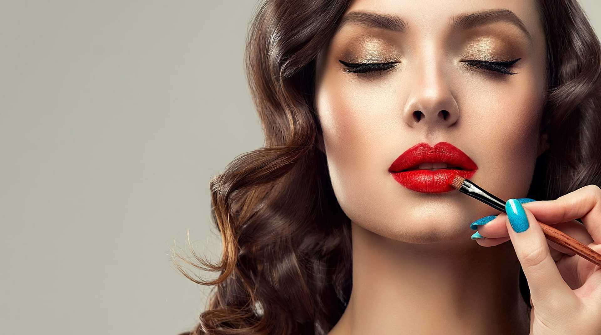 Woman's lips being applied with red lipstick by a hand with blue nails.