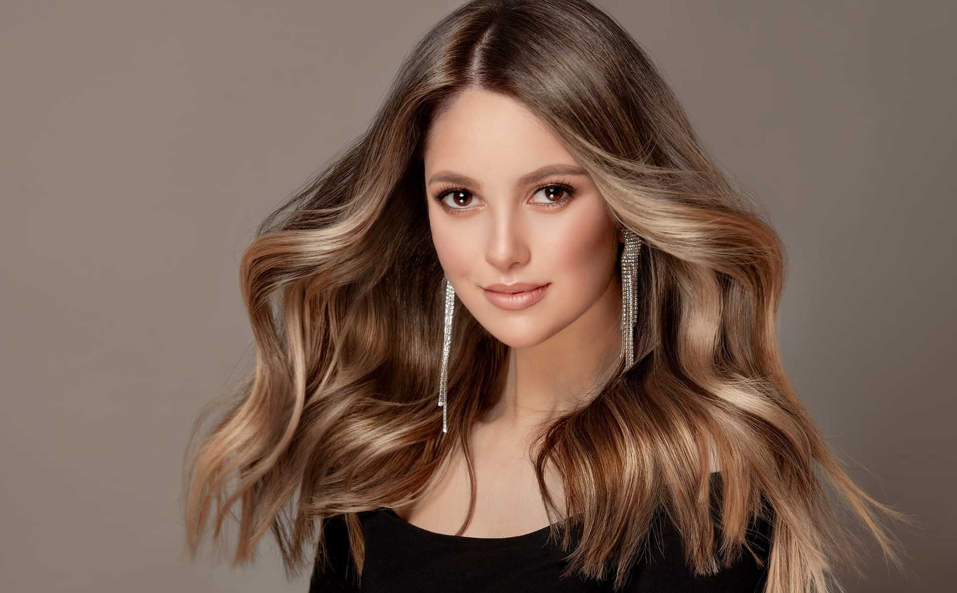 Young woman with flowing hair and long earrings, posing against a neutral background.